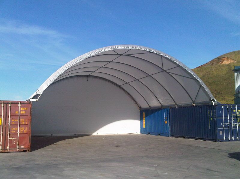Full Endwall Fabric Shelter A fabric shelter with a full endwall design, supported by containers for industrial or storage use.